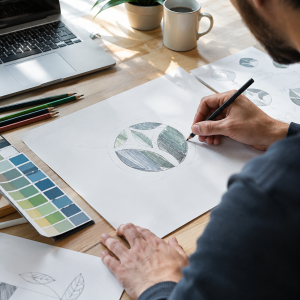 Immersion dans le processus de création de logo artisanal et numérique par Blean Studio. Ce visuel capture l'art de l'esquisse manuelle sur un bureau en bois chaleureux. Un designer professionnel affine minutieusement les détails d'un logotype circulaire aux motifs organiques et naturels, remplissant les formes au crayon. Autour de lui, on aperçoit des outils de design essentiels : un nuancier Pantone, des crayons de couleur, un ordinateur portable et une tasse de café, illustrant la fusion entre l'artisanat traditionnel et la technologie dans l'élaboration de l'identité visuelle d'une marque.